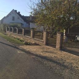 MDJ Metalltechnik Dawid Janczyk - Nowoczesne ogrodzenie z elementami kutymi i kamiennymi słupkami, brama wjazdowa z metalowymi prętami, widok z ulicy na tle domu i drzew