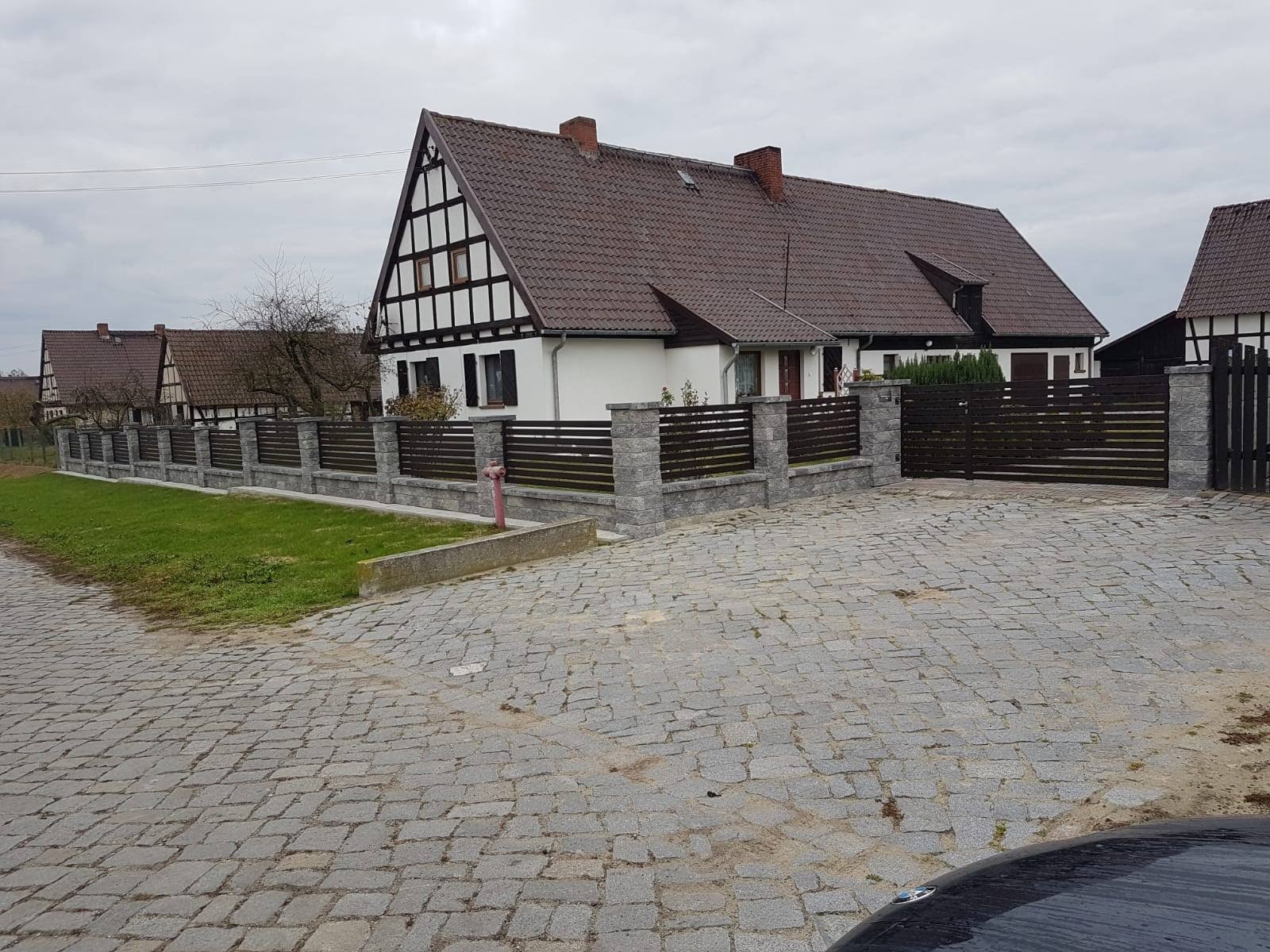 Nowoczesne ogrodzenie posesji z drewnianymi panelami poziomymi i kamiennymi słupkami, uzupełnione bramą wjazdową, na tle domu w stylu szachulcowym pod pochmurnym niebem.