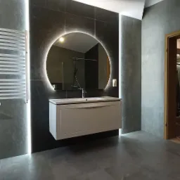 Nowoczesna łazienka w Powałkach z szarymi płytkami, okrągłym lustrem z podświetleniem LED i wiszącą szafką umywalkową. Widoczny także grzejnik drabinkowy i fragment otwartych drzwi.