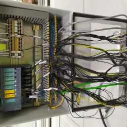 Otwarta szafa sterownicza z widocznymi modułami Siemens SIMATIC ET 200SP, listwami zaciskowymi i gąszczem przewodów oznaczonych etykietami, przewody czarne, żółto-zielone i niebieskie, perspektywa...