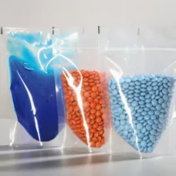 Trzy przezroczyste torebki strunowe typu doypack wypełnione: niebieską cieczą, pomarańczowymi drażetkami i jasnoniebieskimi drażetkami, ustawione na białym tle.