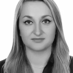 Portret kobiety z długimi blond włosami, ubranej w ciemną koszulę i delikatny naszyjnik z krzyżykiem, na jasnym tle. Czarno-biała fotografia.