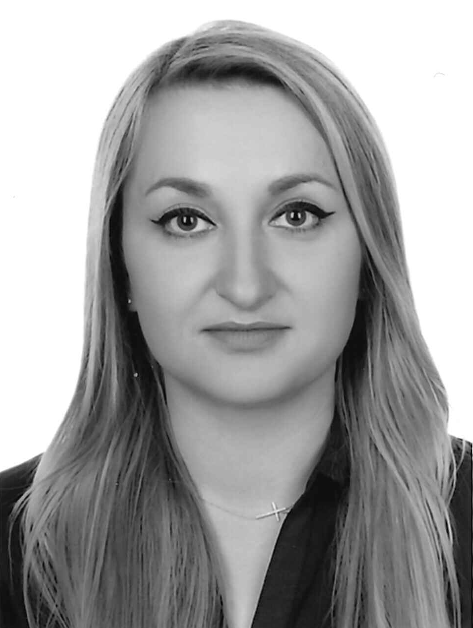 Portret kobiety z długimi blond włosami, ubranej w ciemną koszulę i delikatny naszyjnik z krzyżykiem, na jasnym tle. Czarno-biała fotografia.