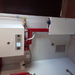 Biały piec grzewczy marki Prestige ACV z czerwonymi rurami i białymi elementami instalacji, zamontowany na kafelkowanej ścianie, widoczne przyłącza i przewody.