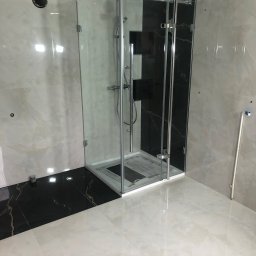 euro-S - Nowoczesna kabina prysznicowa z transparentnego szkła, chromowane okucia, czarne i beżowe płytki imitujące marmur na ścianach i podłodze, widoczne rury instalacyjne.