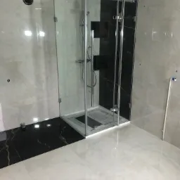 Nowoczesna kabina prysznicowa z transparentnego szkła, chromowane okucia, czarne i beżowe płytki imitujące marmur na ścianach i podłodze, widoczne rury instalacyjne.