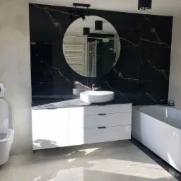 Nowoczesna łazienka z czarnymi płytkami imitującymi marmur, okrągłe lustro z podświetleniem i biała szafka z umywalką nablatową oraz wolnostojąca wanna, oświetlona promieniami słonecznymi.