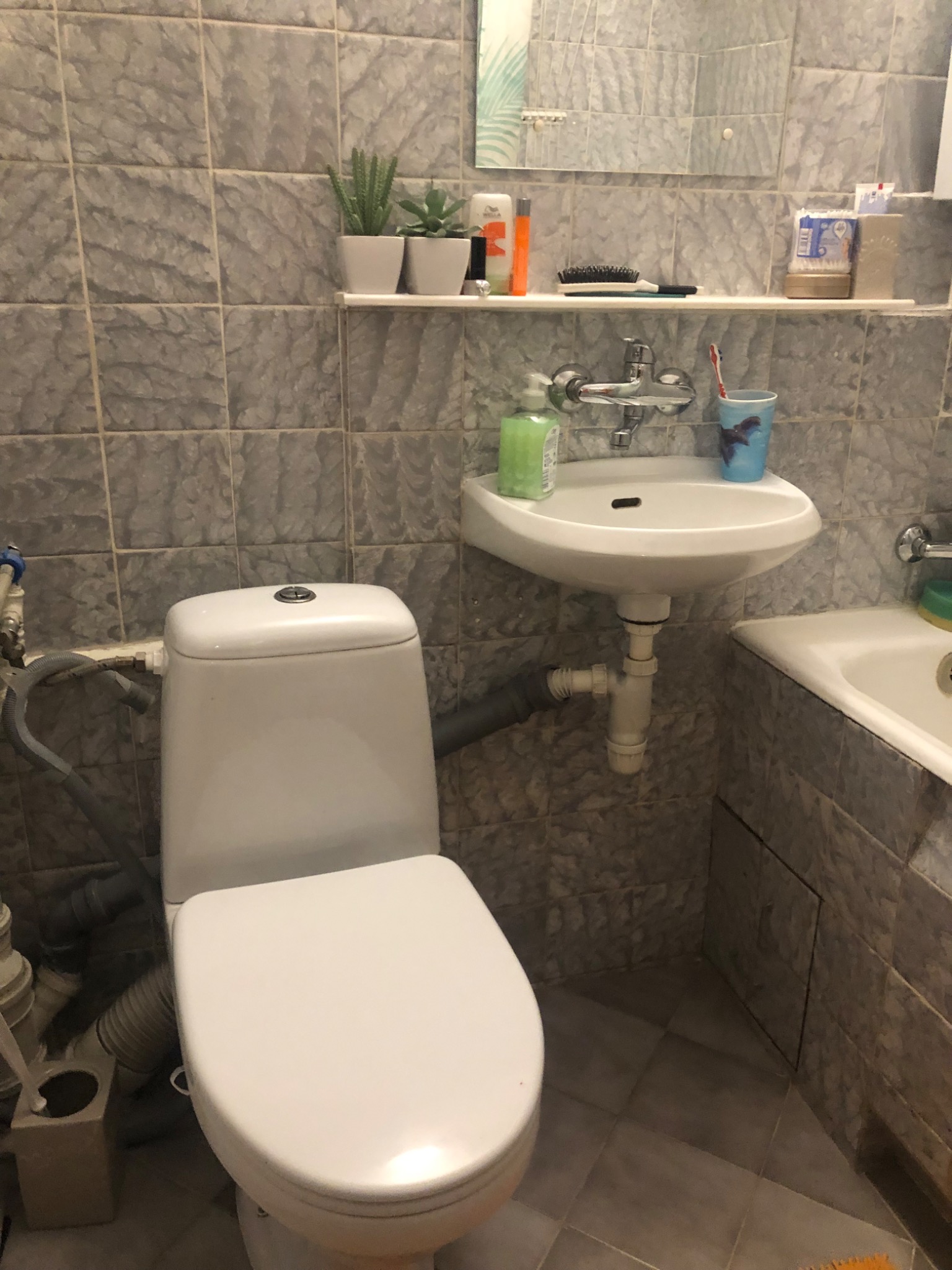 Ujęcie standardowej łazienki z szarymi płytkami, umywalką nablatową, toaletą i wanną, z widoczną instalacją hydrauliczną i akcesoriami łazienkowymi na półce.