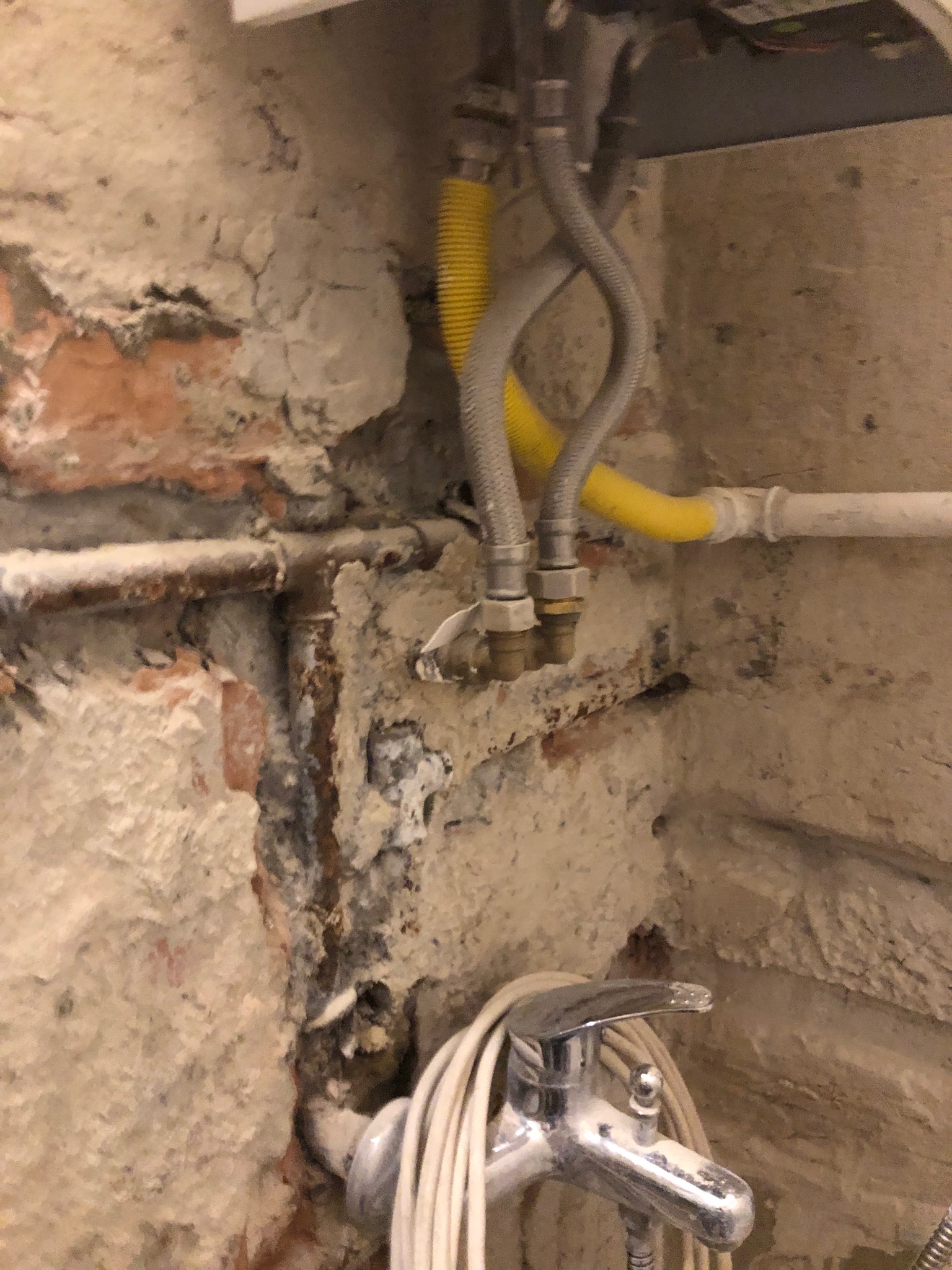 Niedokończona instalacja wodno-kanalizacyjna w łazience z odsłoniętą cegłą i fragmentem pieca gazowego, widoczne rury miedziane, żółta rura gazowa i bateria umywalkowa.