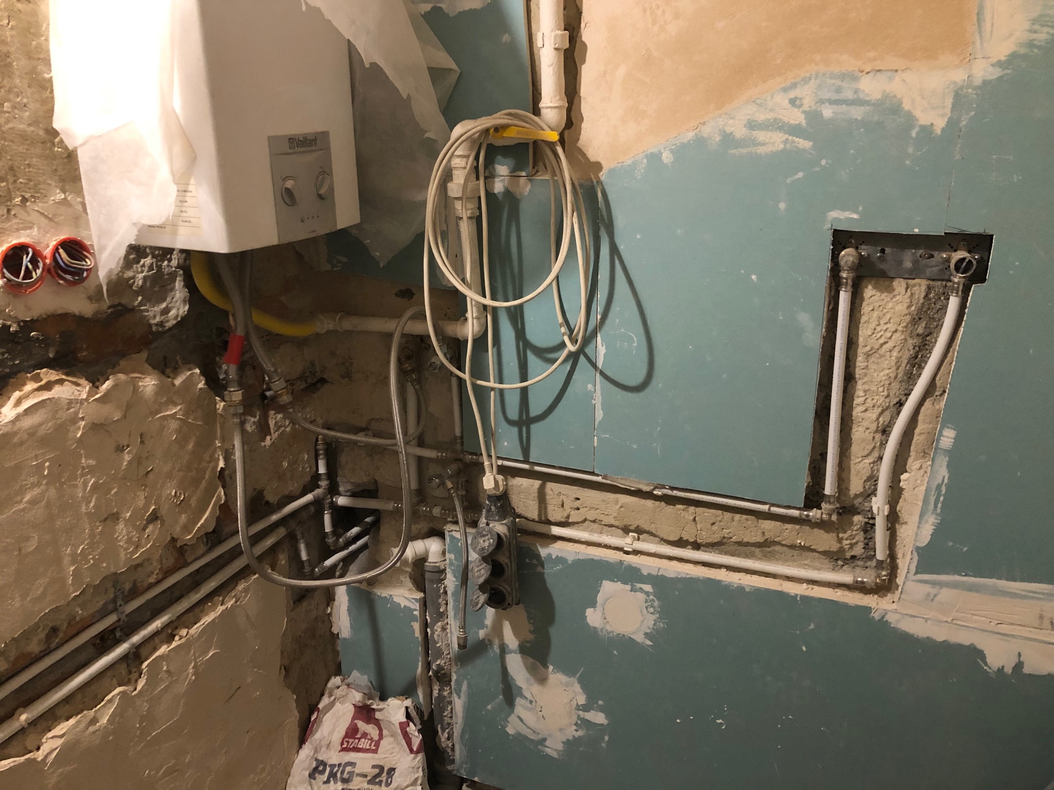Ściana w trakcie remontu z widoczną instalacją hydrauliczną, piecem gazowym Vaillant i workiem zaprawy Stabili PKG-25, ukazująca fragmenty uszkodzonego tynku i przygotowania do dalszych prac...
