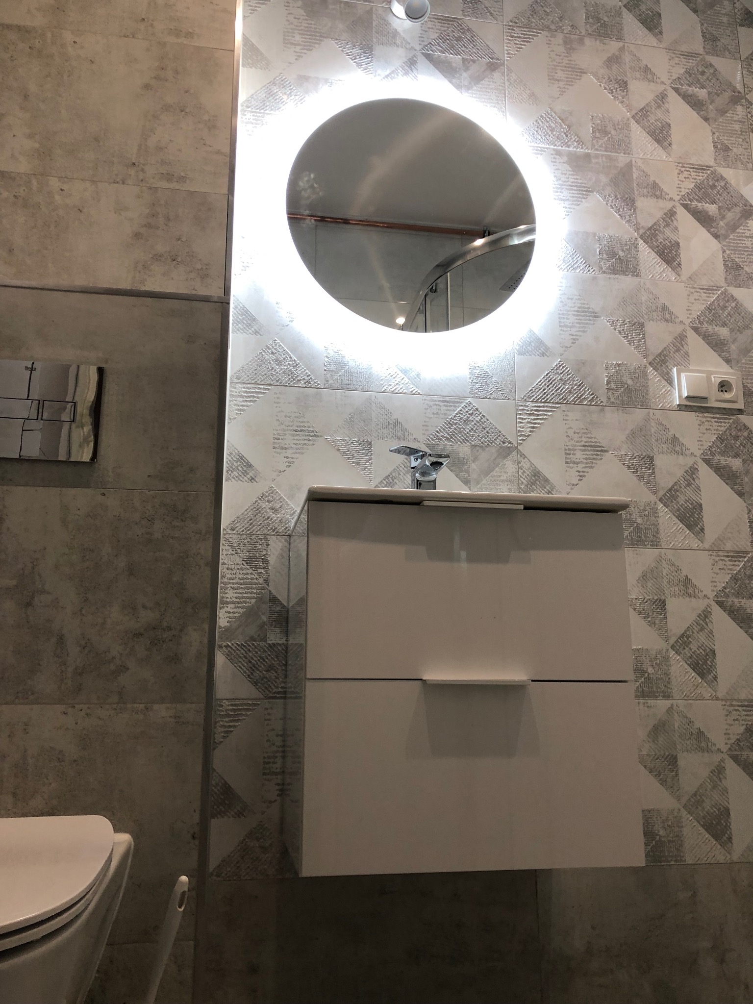 Nowoczesna łazienka: biała szafka pod umywalkę z dwoma szufladami, okrągłe lustro z podświetleniem LED, bateria umywalkowa, wzorzyste szaro-białe płytki na ścianie, wisząca toaleta z chromowanym...