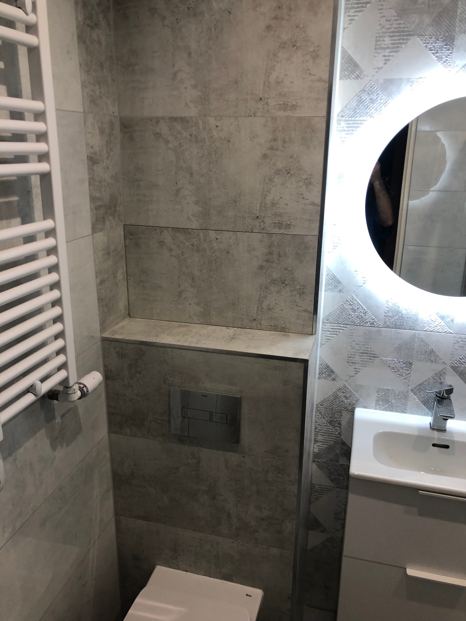 Szara łazienka z podświetlanym lustrem, grzejnikiem drabinkowym i zabudowaną spłuczką WC z ukrytym schowkiem.