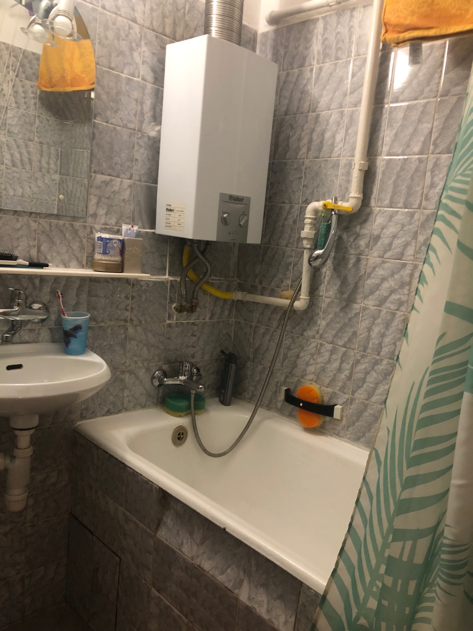 Standardowa łazienka z szarymi płytkami, białą wanną i umywalką, bojlerem gazowym na ścianie, widok zza zasłony prysznicowej.