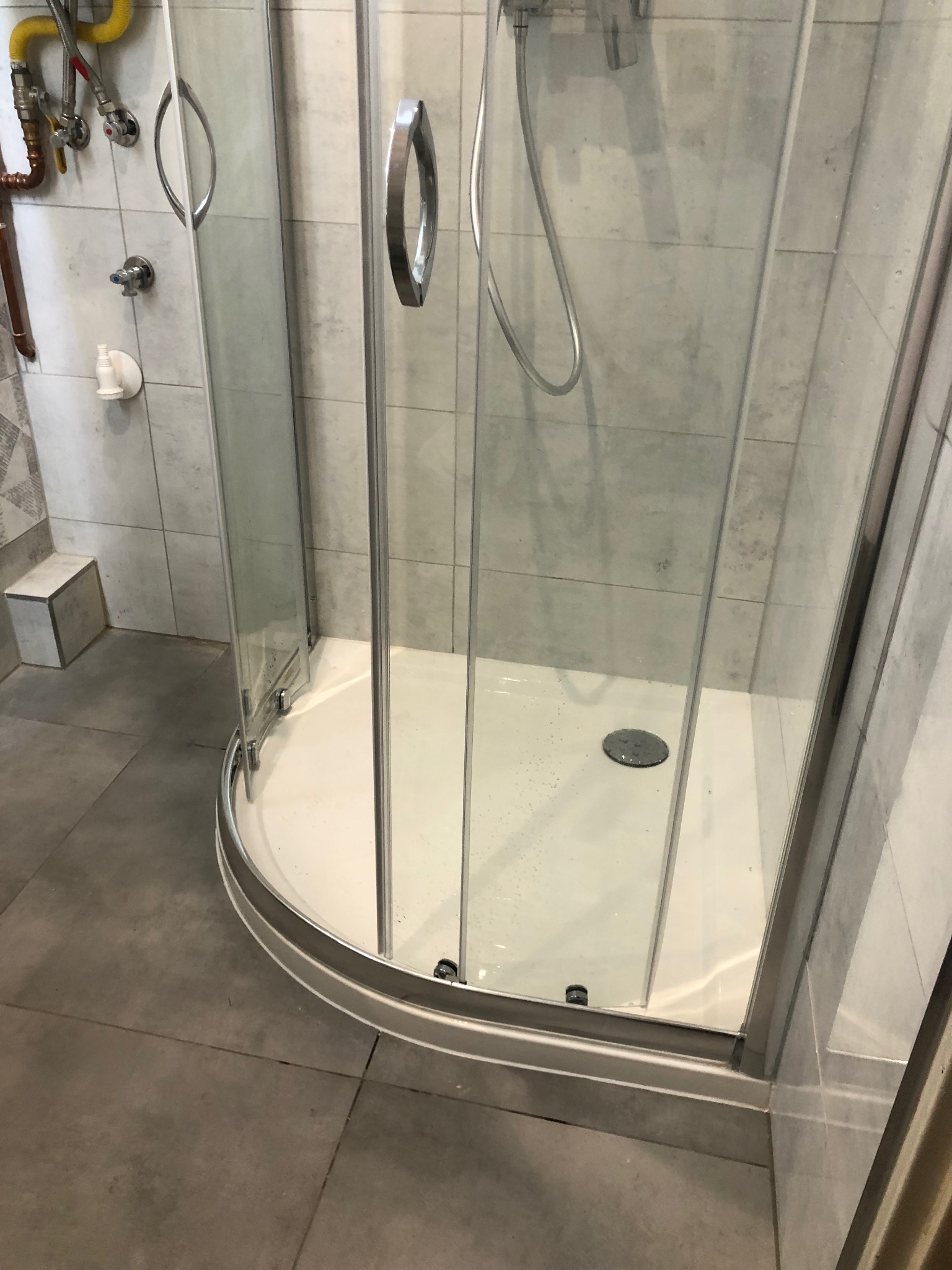 Narożna kabina prysznicowa z przezroczystymi drzwiami i srebrnymi uchwytami, szare płytki na ścianach i podłodze, widoczne rury instalacji wodnej w rogu.
