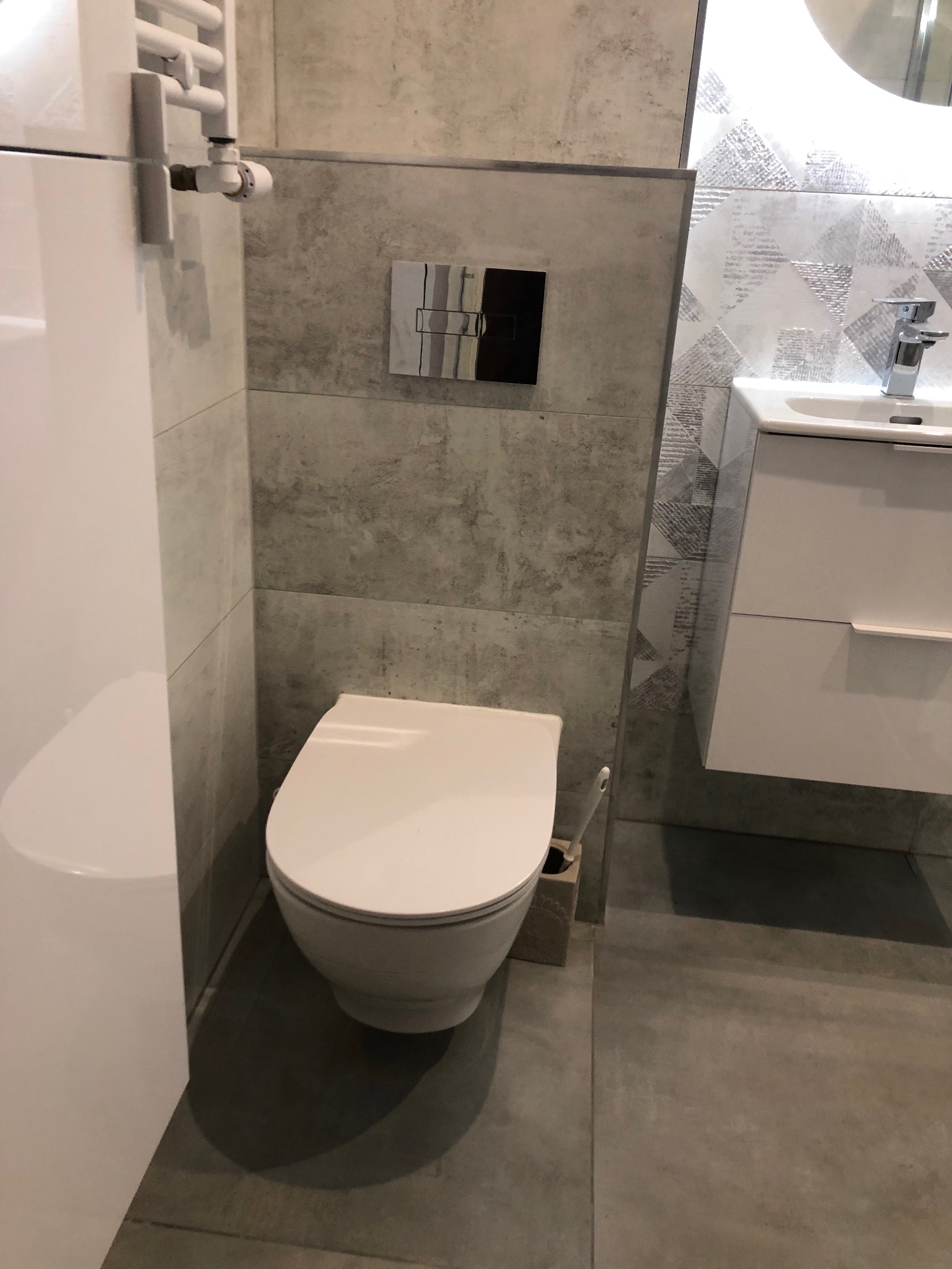 Nowoczesna łazienka z podwieszaną toaletą, umywalką z szafką i grzejnikiem drabinkowym, szara glazura na ścianach i podłodze, dekoracyjne płytki za umywalką.