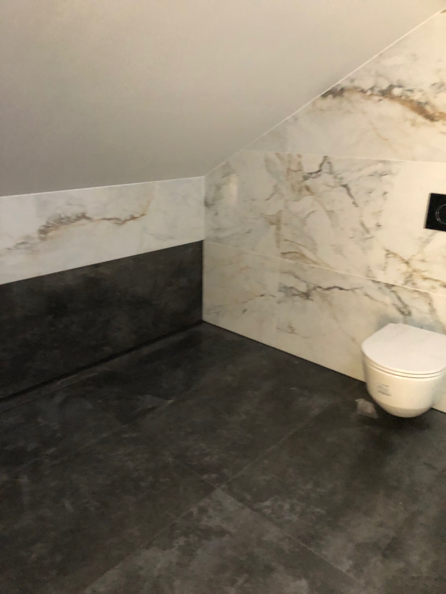 Nowoczesna łazienka na poddaszu z marmurowymi płytkami i podwieszaną toaletą, czarne płytki podłogowe i biało-szare na ścianach, skośny sufit.