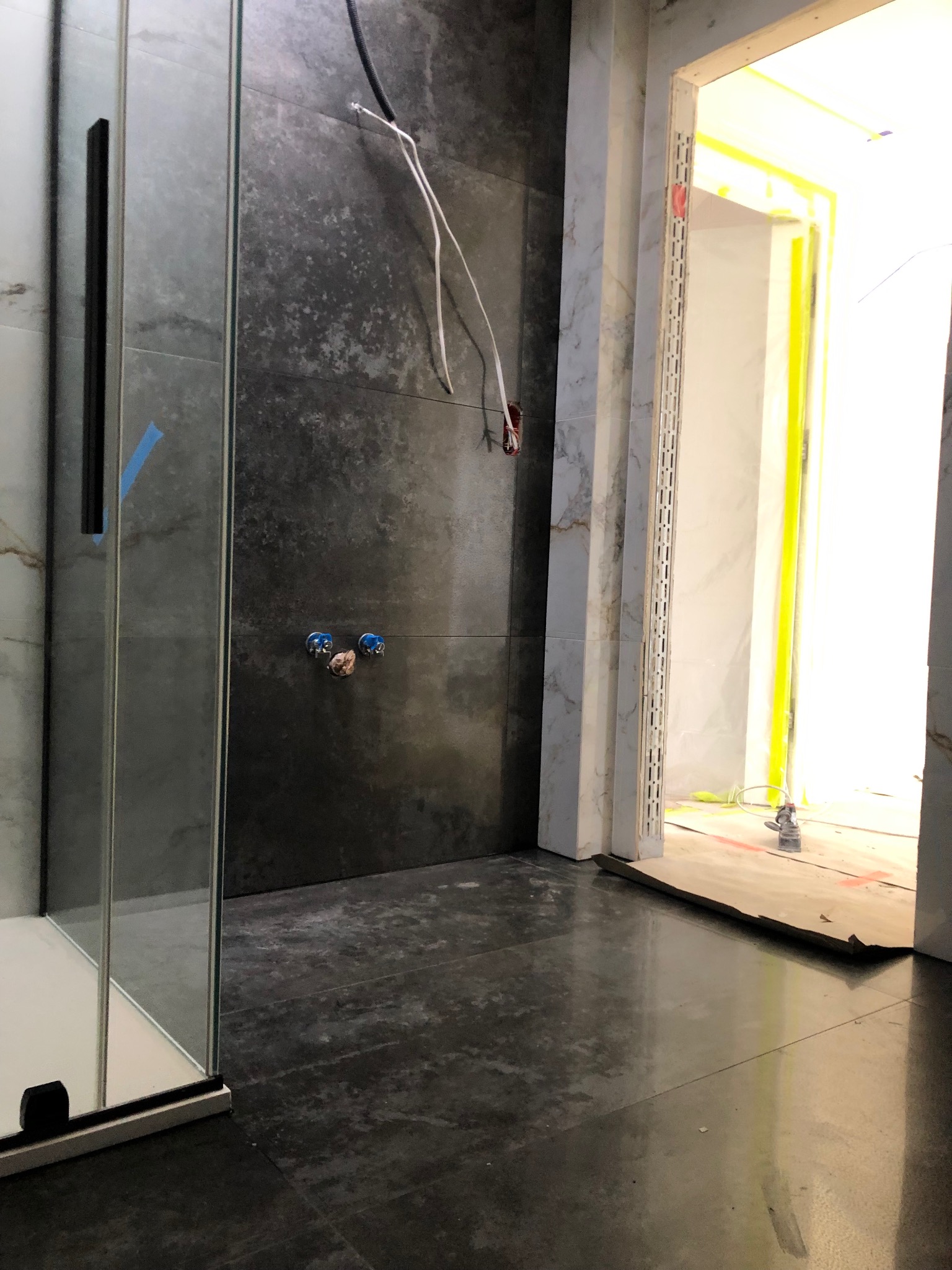 Wnętrze łazienki w trakcie prac wykończeniowych: widoczna kabina prysznicowa z niebieską taśmą zabezpieczającą, ciemnoszare płytki na ścianach z wystającymi przewodami i przygotowanymi punktami...
