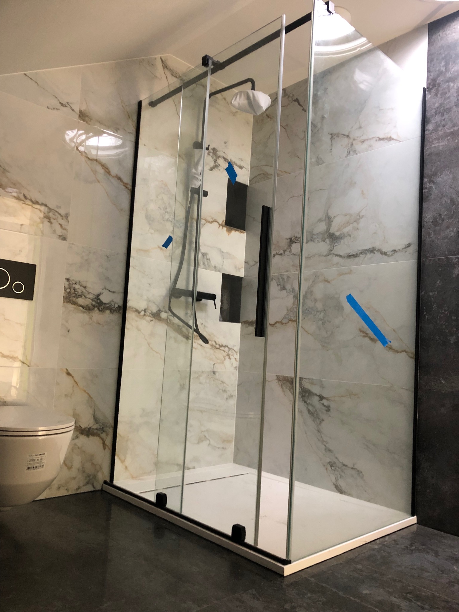 Nowoczesna kabina prysznicowa z czarnymi profilami, marmurowe płytki na ścianach, widoczne niebieskie taśmy zabezpieczające, częściowo widoczna toaleta.