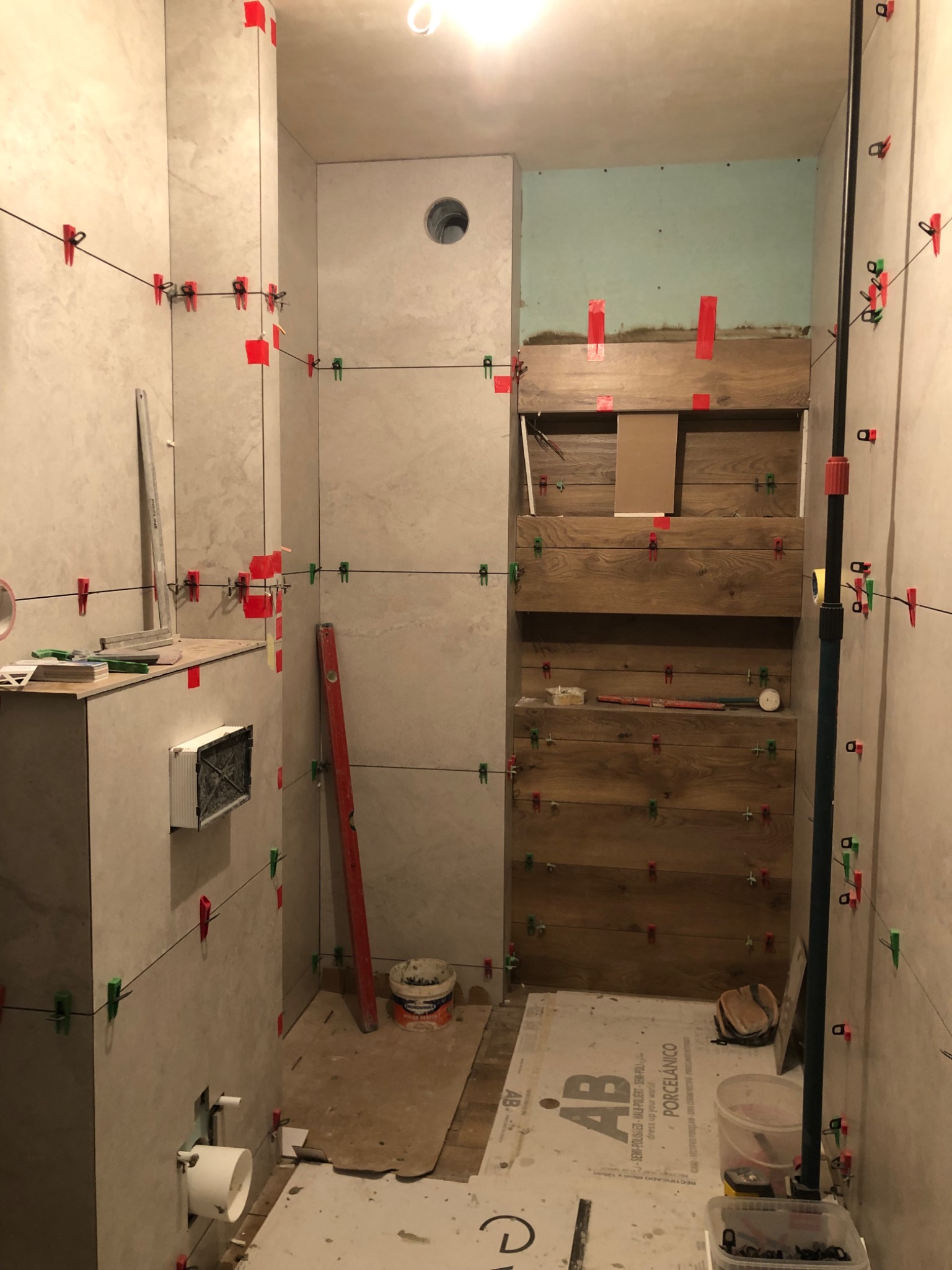 Wnętrze łazienki w trakcie prac wykończeniowych, częściowo wyłożone jasnymi płytkami, z elementami drewnopodobnymi na jednej ze ścian. Widoczne narzędzia glazurnicze, poziomica, krzyżyki dystansowe...