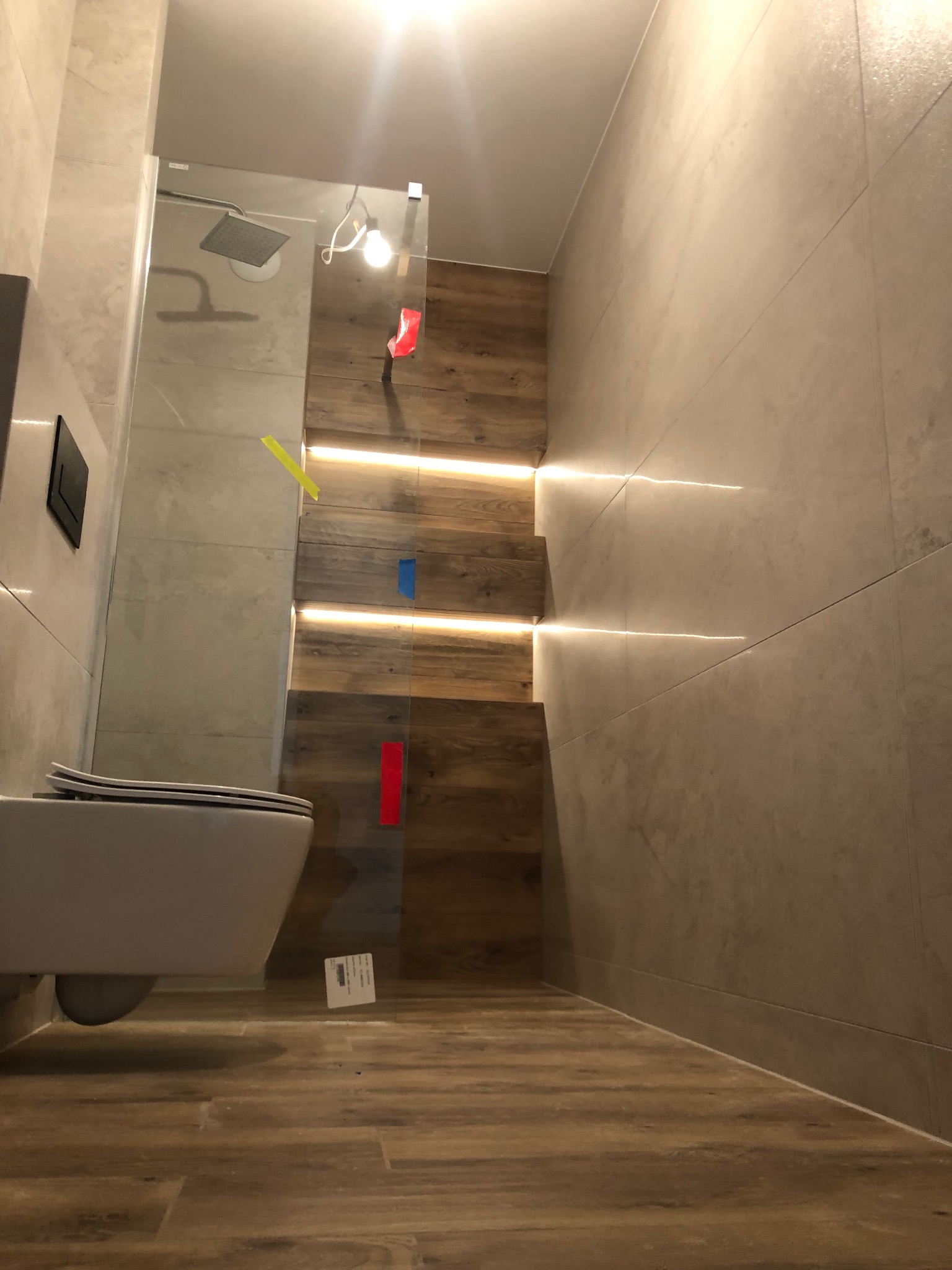 Nowoczesna łazienka w trakcie wykańczania, widoczne płytki imitujące drewno na podłodze i ścianie, szklana kabina prysznicowa z deszczownicą, podwieszana miska WC, taśmy malarskie na szkle...