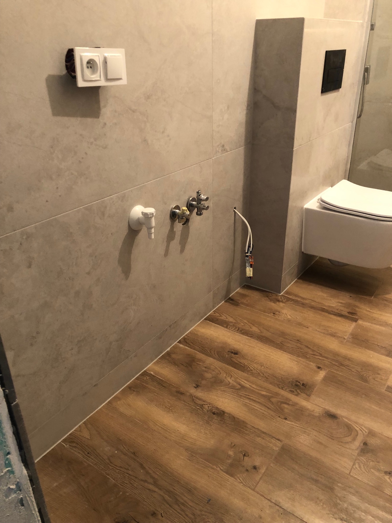 Fragment łazienki w trakcie prac wykończeniowych: płytki imitujące marmur na ścianach, drewnopodobna podłoga, wisząca toaleta, wystające kable i rury wodne, zamontowane gniazdko elektryczne...