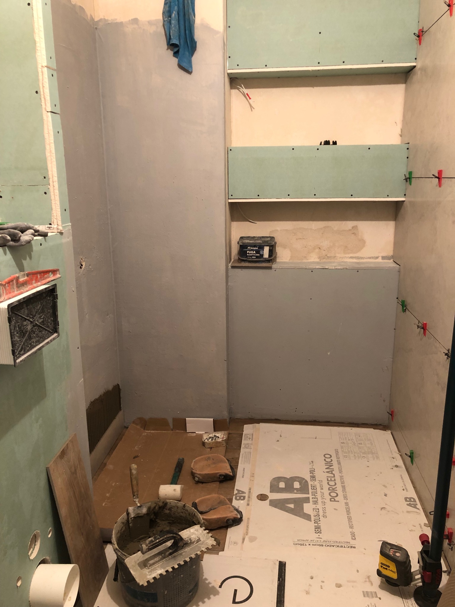 Łazienka w trakcie remontu: przygotowane ściany z płyt gipsowo-kartonowych, narzędzia glazurnicze, wiadro z zaprawą, płytki podłogowe AB Porcelanico i poziomica laserowa na podłodze.