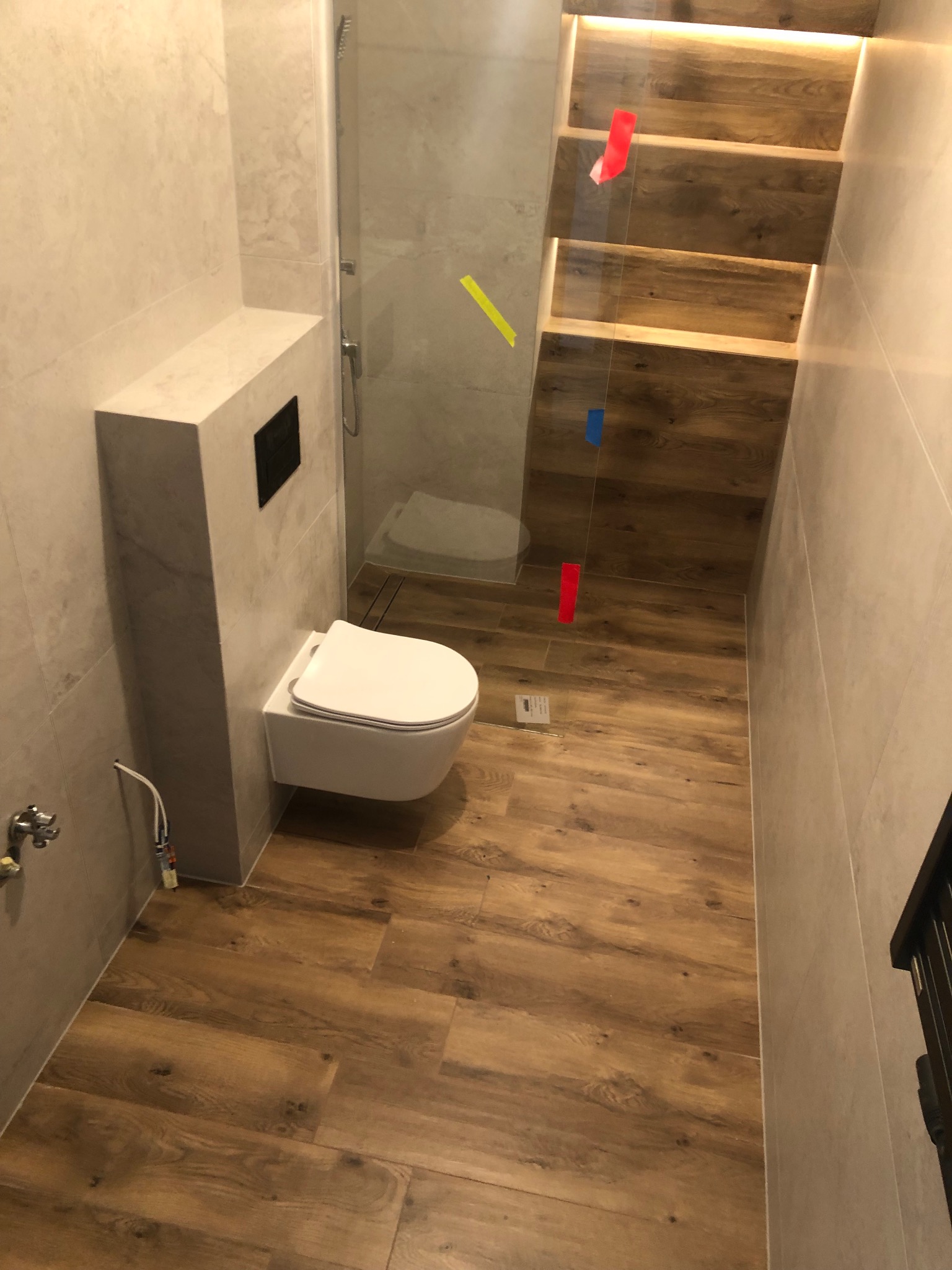 Nowoczesna łazienka z wiszącą toaletą, płytkami imitującymi drewno na podłodze oraz podświetlanymi półkami w ścianie, zabezpieczonymi taśmą malarską.