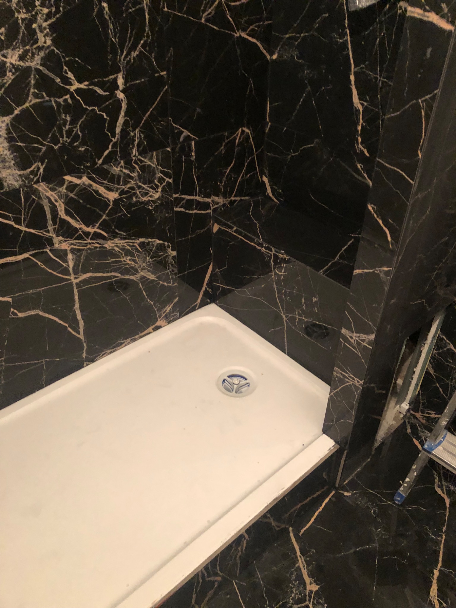 Biały brodzik w narożniku łazienki wyłożonej czarnym marmurem z złotymi żyłami, widoczny fragment drabiny malarskiej.