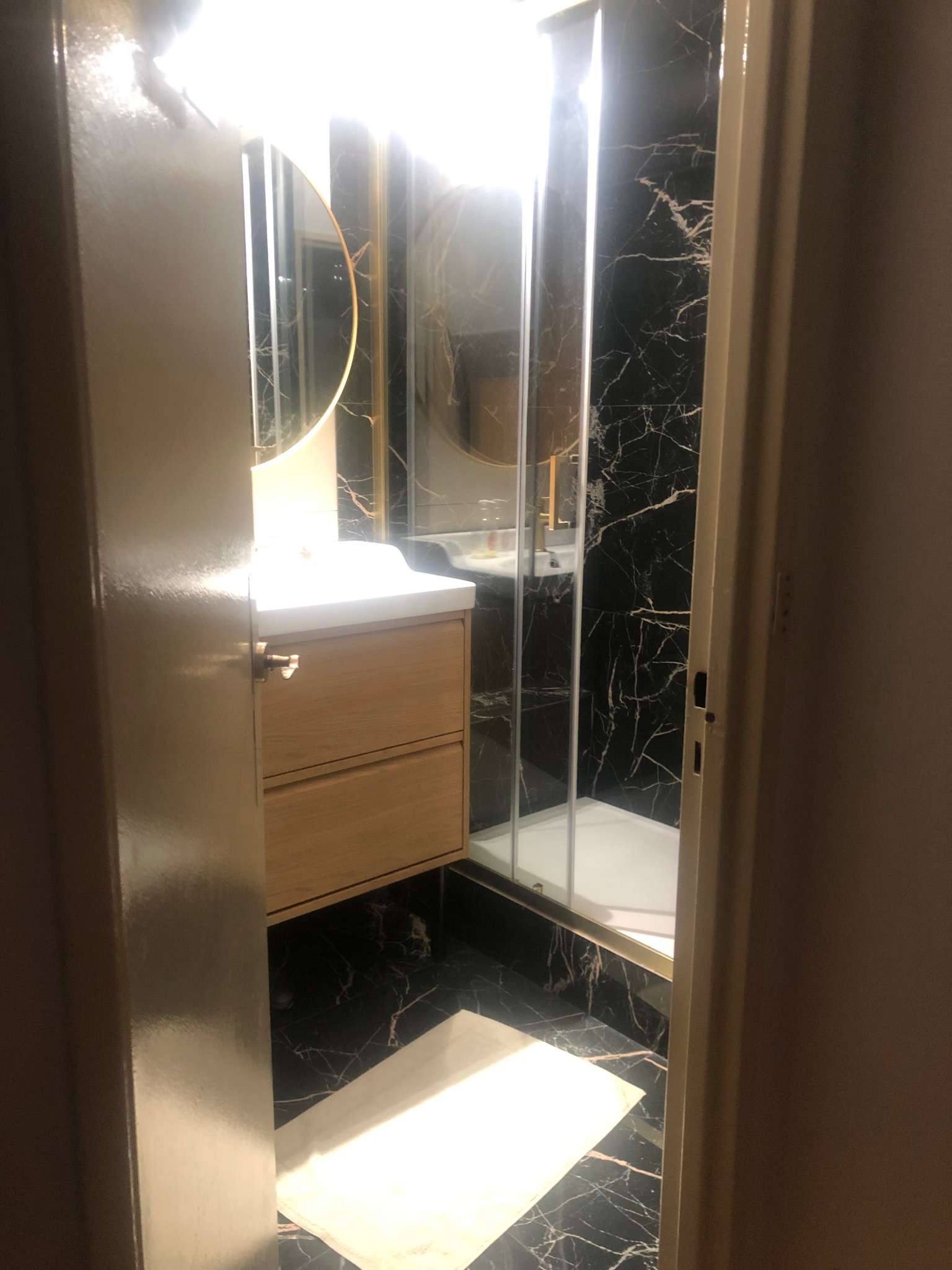 Nowoczesna łazienka z czarnymi marmurowymi ścianami, kabiną prysznicową ze złotymi detalami i umywalką z szafką z jasnego drewna, widziana zza uchylonych drzwi.