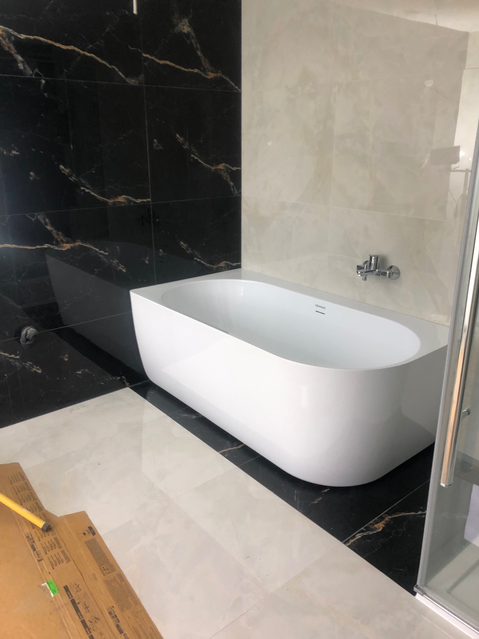 Nowoczesna, wolnostojąca wanna na tle marmurowych płytek w czarno-złotej kolorystyce, z elementami szklanej kabiny prysznicowej i fragmentem kartonu na podłodze.