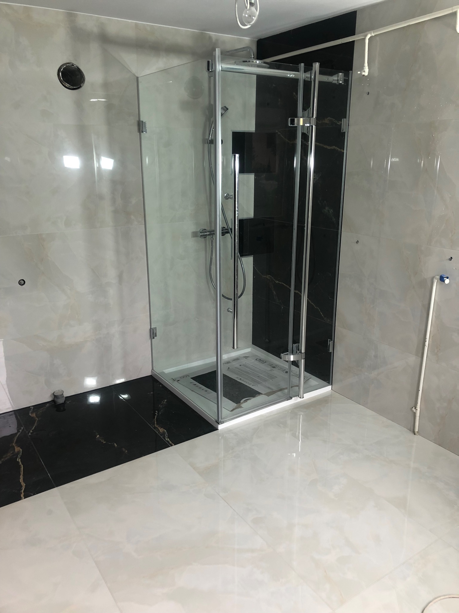 Nowoczesna kabina prysznicowa z transparentnego szkła, chromowane okucia, czarne i beżowe płytki imitujące marmur na ścianach i podłodze, widoczne rury instalacyjne.