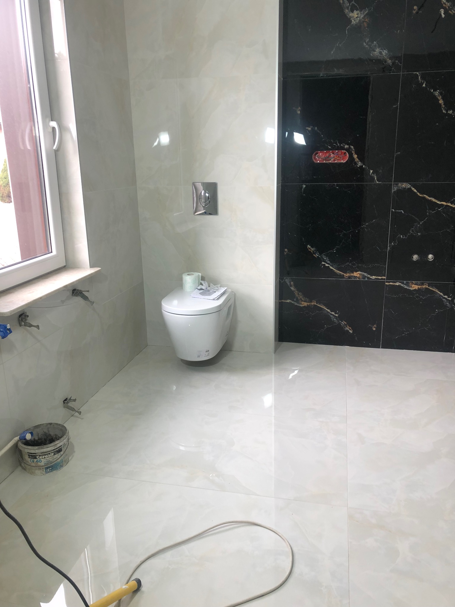 Nowoczesna łazienka w trakcie wykańczania: białe i czarne marmurowe płytki, wisząca toaleta, widoczne narzędzia i wiadro z zaprawą.