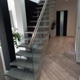 Nowoczesne schody wspornikowe z szarymi stopniami, szklaną balustradą i metalową poręczą w minimalistycznym wnętrzu z marmurową podłogą i dyskretnym oświetleniem punktowym.