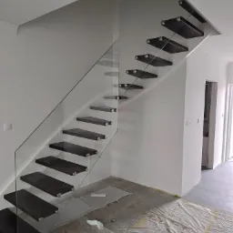 Nowoczesne schody wspornikowe z ciemnego drewna, szklana balustrada, mocowane do ściany za pomocą metalowych elementów, zabezpieczone folią malarską na podłodze.