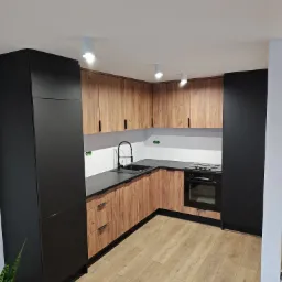 Nowoczesna kuchnia na wymiar z frontami w kolorze drewna i czarnymi szafkami, czarny zlew i bateria, jasna podłoga, oświetlenie punktowe.