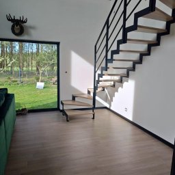 Nowoczesne schody z drewnianymi stopniami i czarną metalową balustradą w jasnym wnętrzu domu z widokiem na zieleń przez duże okno.