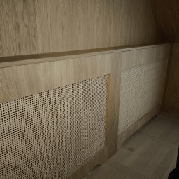 Drewniana zabudowa grzejnika z rattanową plecionką w jasnym odcieniu drewna, dopasowana do boazerii i podłogi. Estetyczne wykończenie wnętrza.