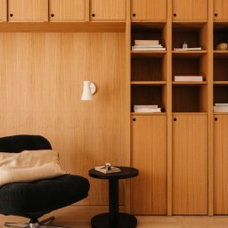 Averit Sławomir Pawłowski - Drewniana zabudowa ścienna z szafkami i półkami, czarny fotel i stolik. Minimalistyczny design, ciepłe oświetlenie, widoczna struktura drewna.