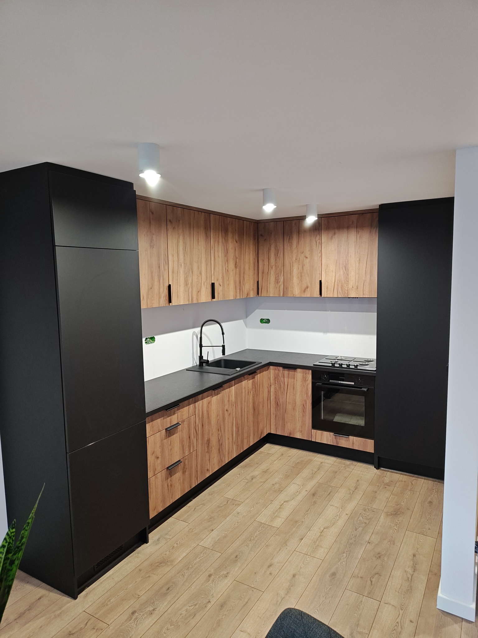 Nowoczesna kuchnia na wymiar z frontami w kolorze drewna i czarnymi szafkami, czarny zlew i bateria, jasna podłoga, oświetlenie punktowe.