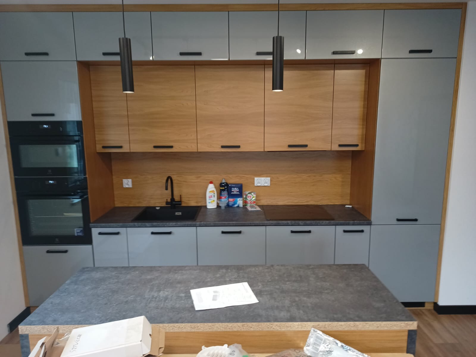 Nowoczesna kuchnia z szarymi i drewnianymi szafkami, czarnym zlewem i płytą indukcyjną. Nad blatem wiszą dwie czarne lampy. Na wyspie kuchennej leżą dokumenty.