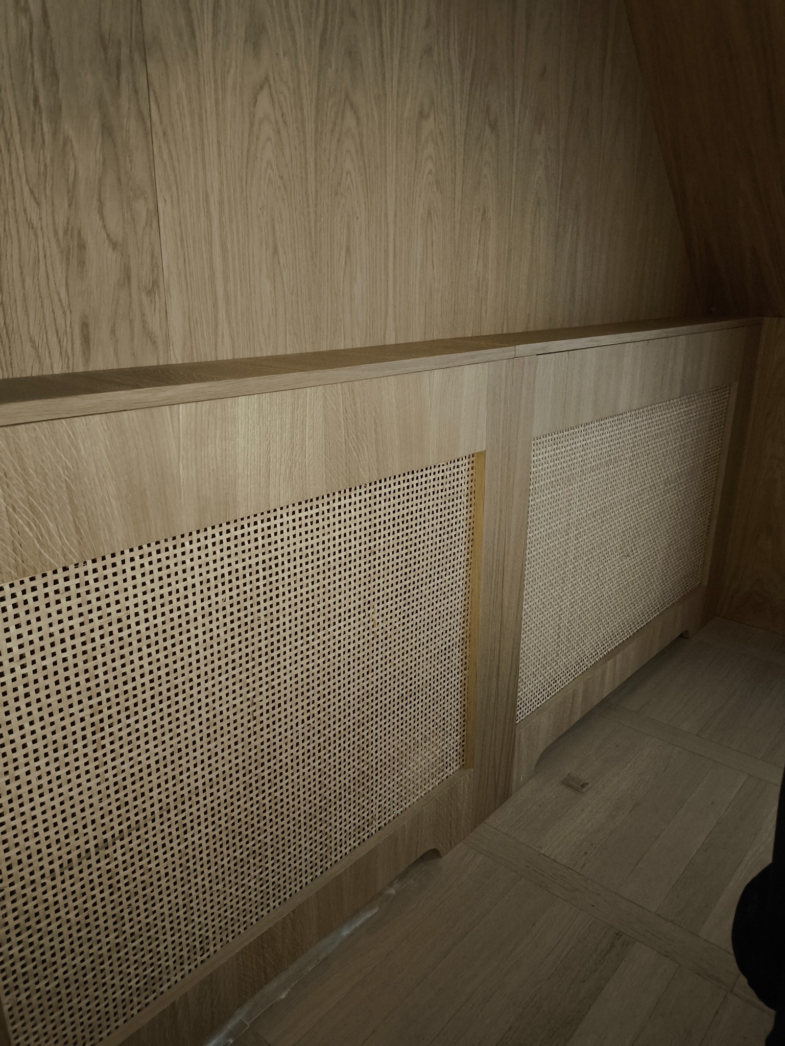 Drewniana zabudowa grzejnika z rattanową plecionką w jasnym odcieniu drewna, dopasowana do boazerii i podłogi. Estetyczne wykończenie wnętrza.