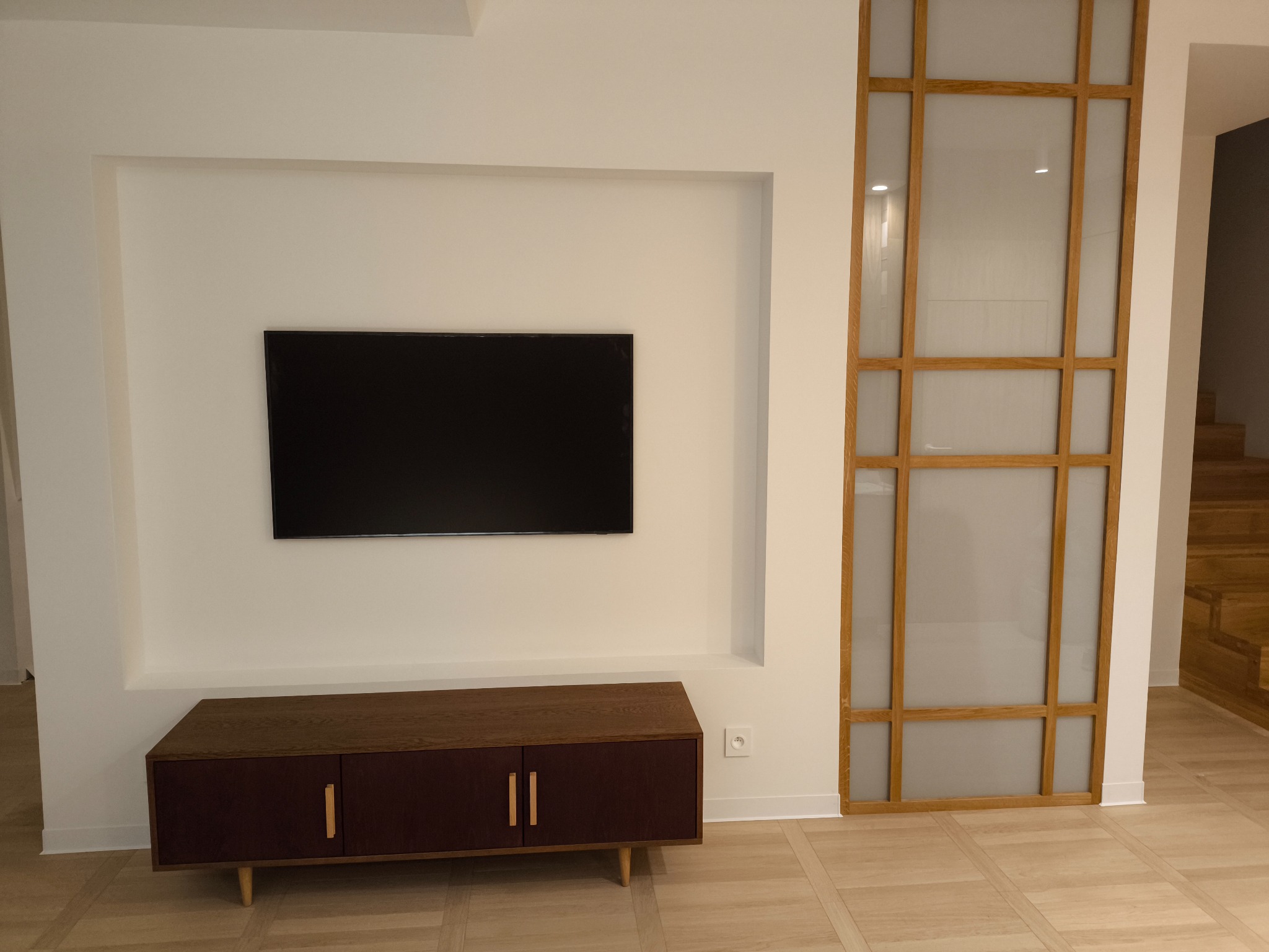 Minimalistyczny salon z drewnianą szafką RTV, telewizorem w białej wnęce i ażurową ścianką działową z drewna, za którą widać fragment drewnianych schodów.