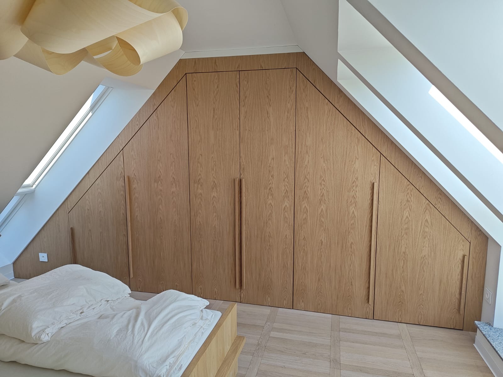 Drewniana szafa wnękowa na poddaszu, dopasowana do skosu dachu. Jasne drewno, minimalistyczne uchwyty, widoczne okno dachowe i fragment łóżka.