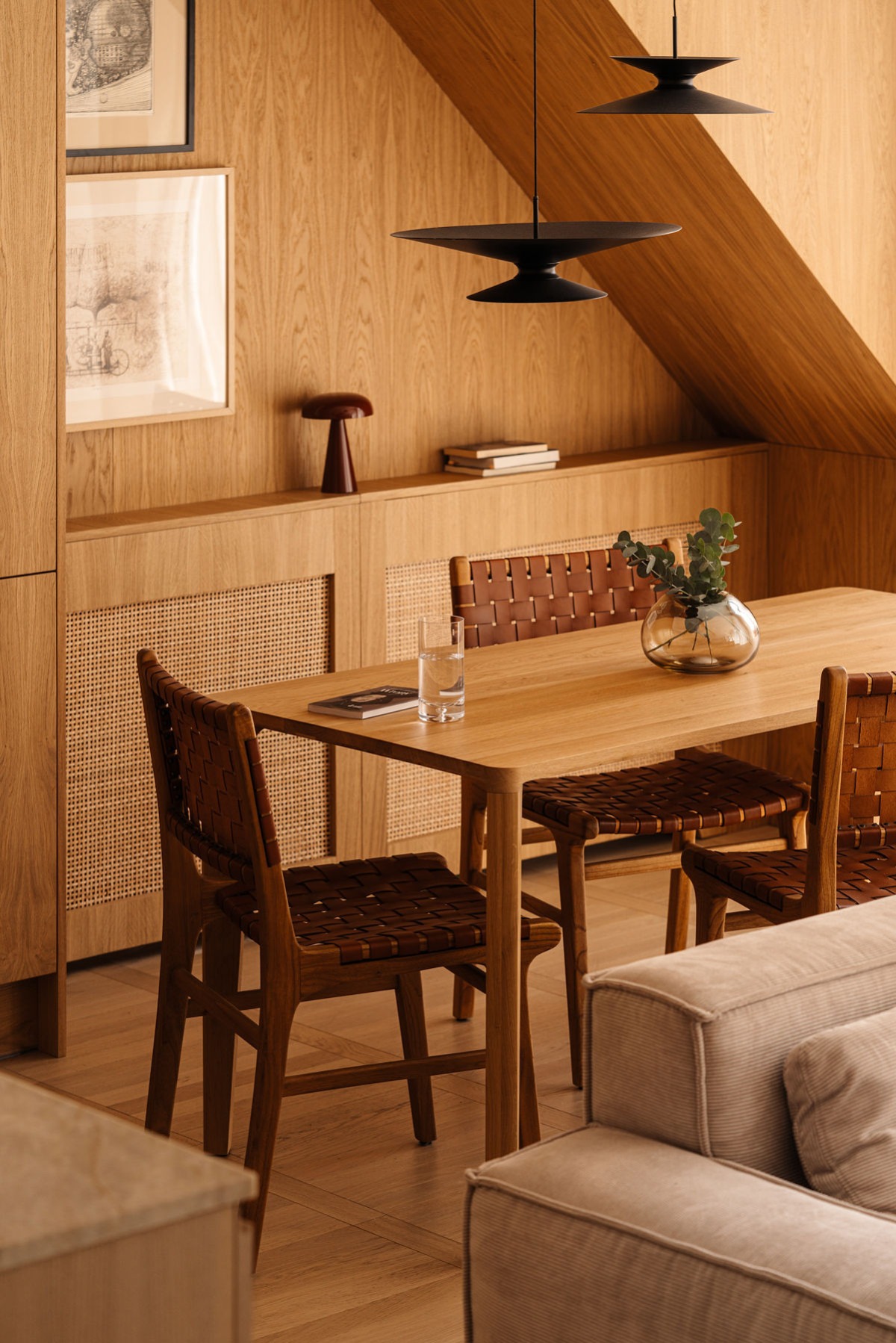 Drewniana zabudowa w skandynawskim stylu z rattanowymi frontami, stół z krzesłami z plecionej skóry, lampy wiszące i skośny sufit na poddaszu.