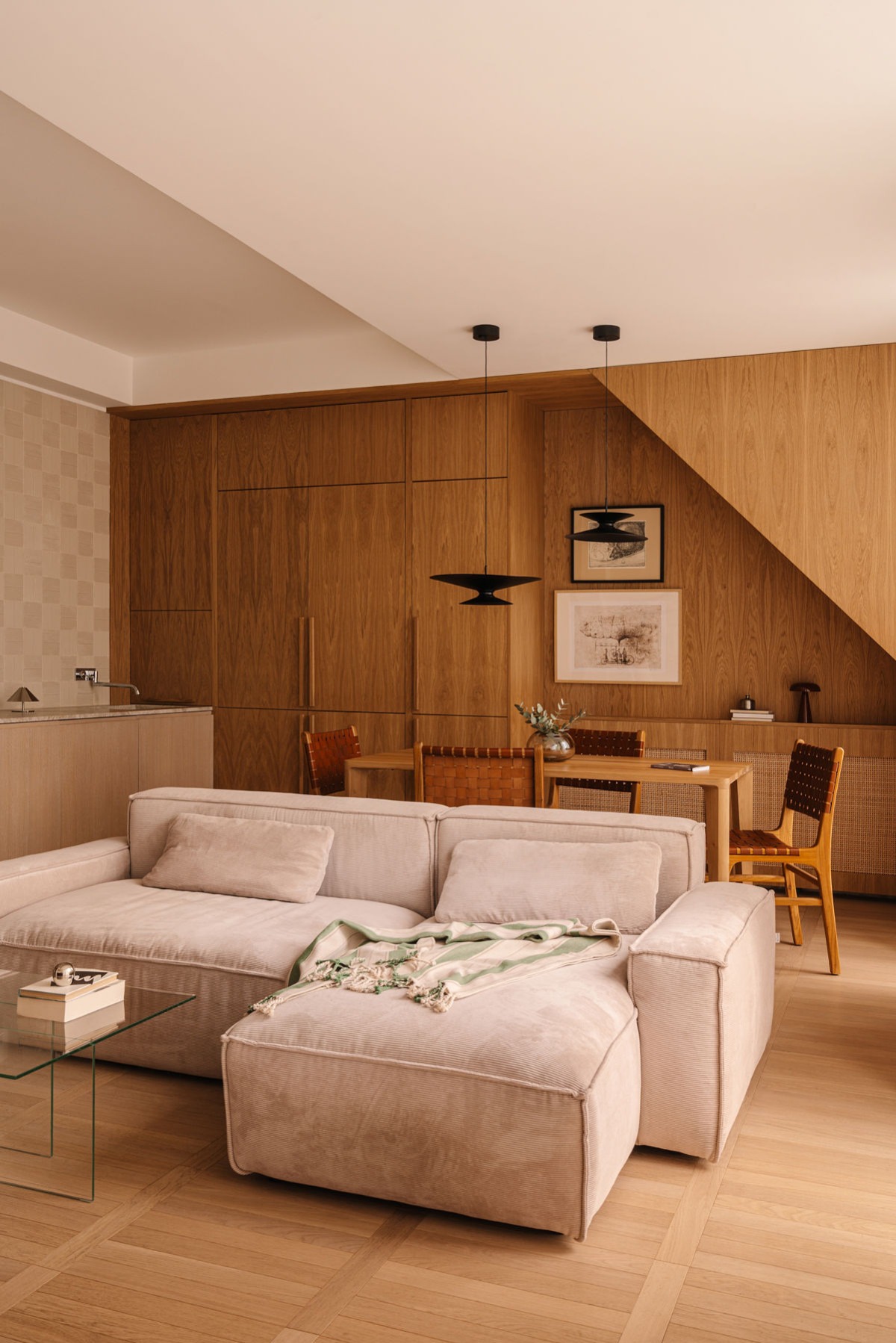 Drewniana zabudowa ściany z szafkami i blatem, wkomponowana w skos dachu. Wnętrze dopełnia sofa, stół z krzesłami i lampy wiszące. Całość utrzymana w ciepłej, naturalnej kolorystyce.