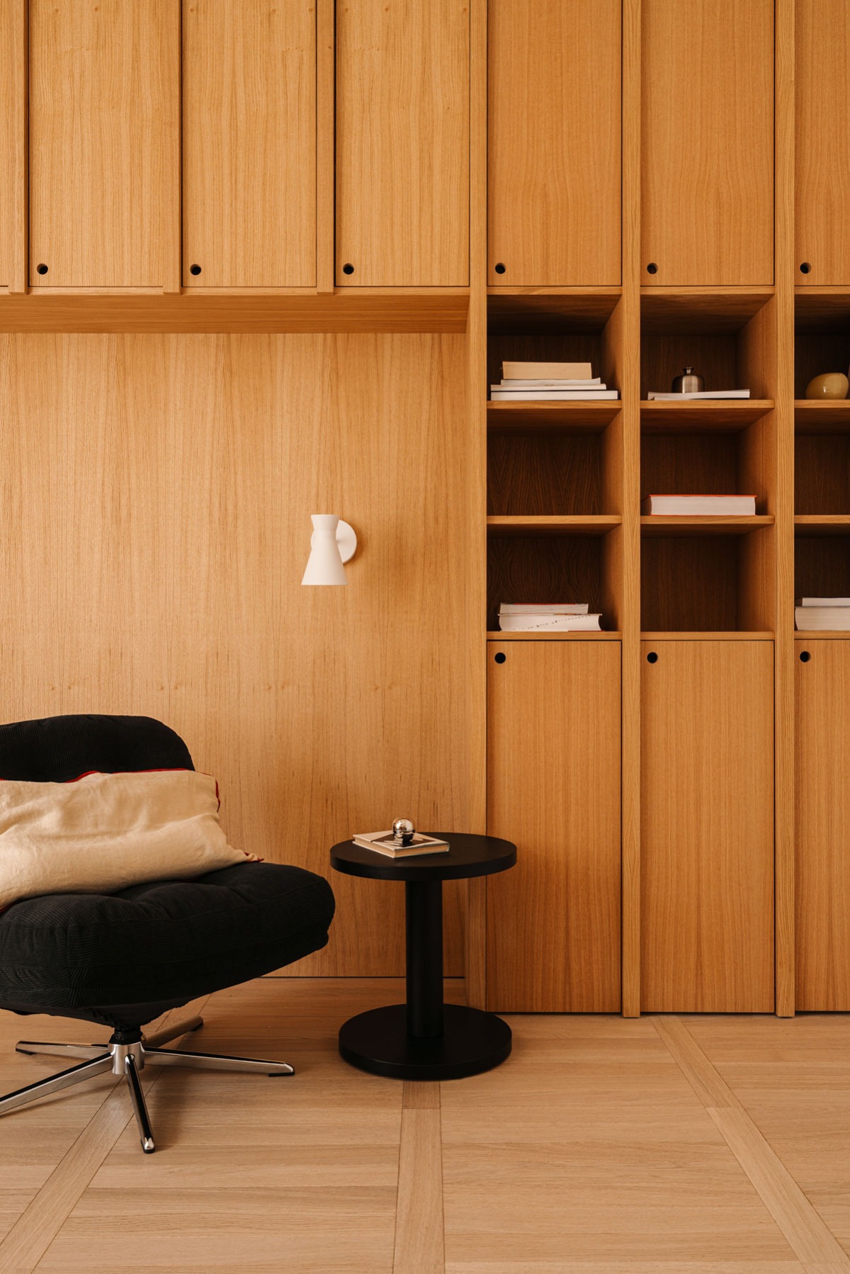 Drewniana zabudowa ścienna z szafkami i półkami, czarny fotel i stolik. Minimalistyczny design, ciepłe oświetlenie, widoczna struktura drewna.