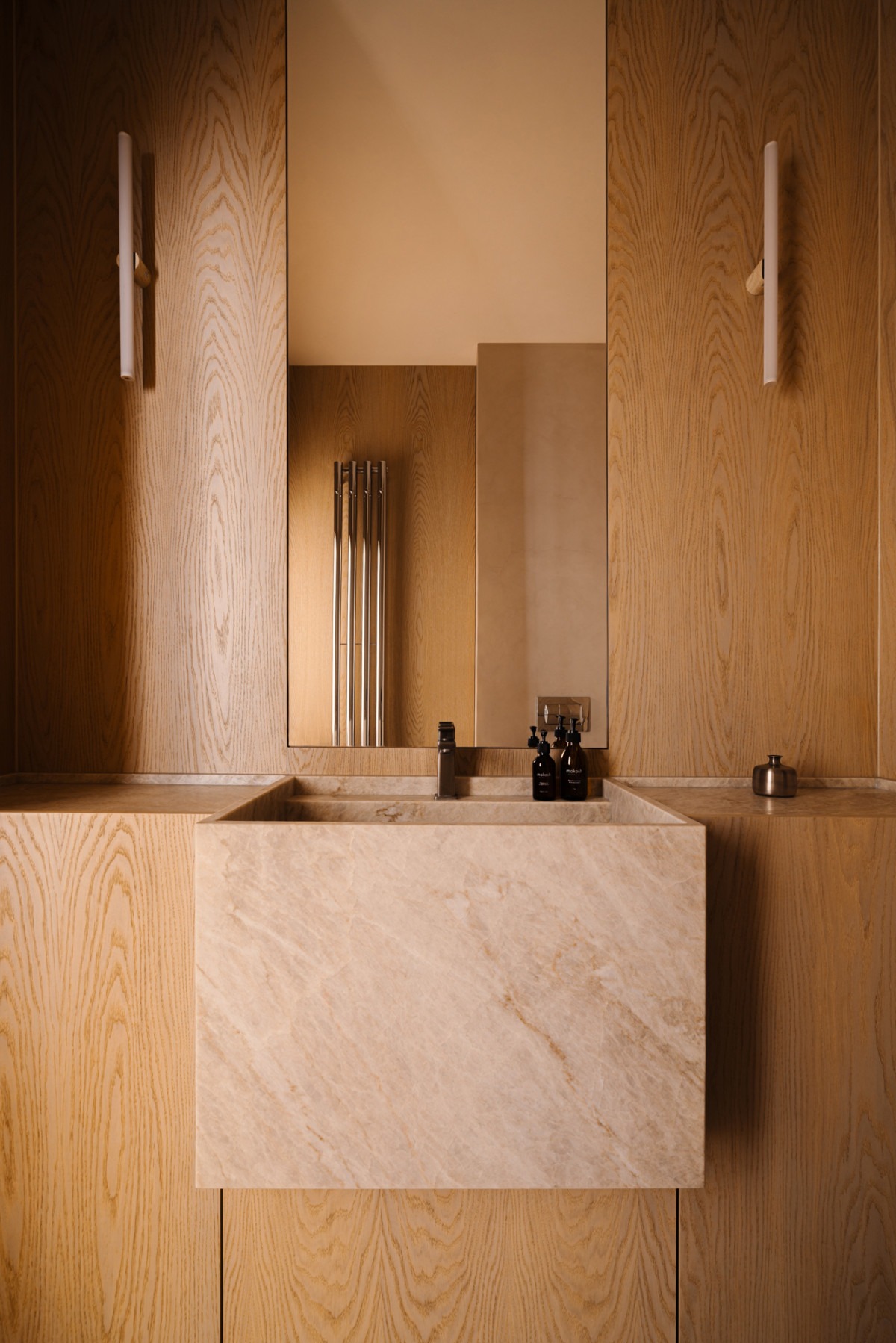 Nowoczesna łazienka z umywalką z beżowego marmuru, drewnianą zabudową i minimalistycznym oświetleniem. W lustrze odbija się grzejnik.