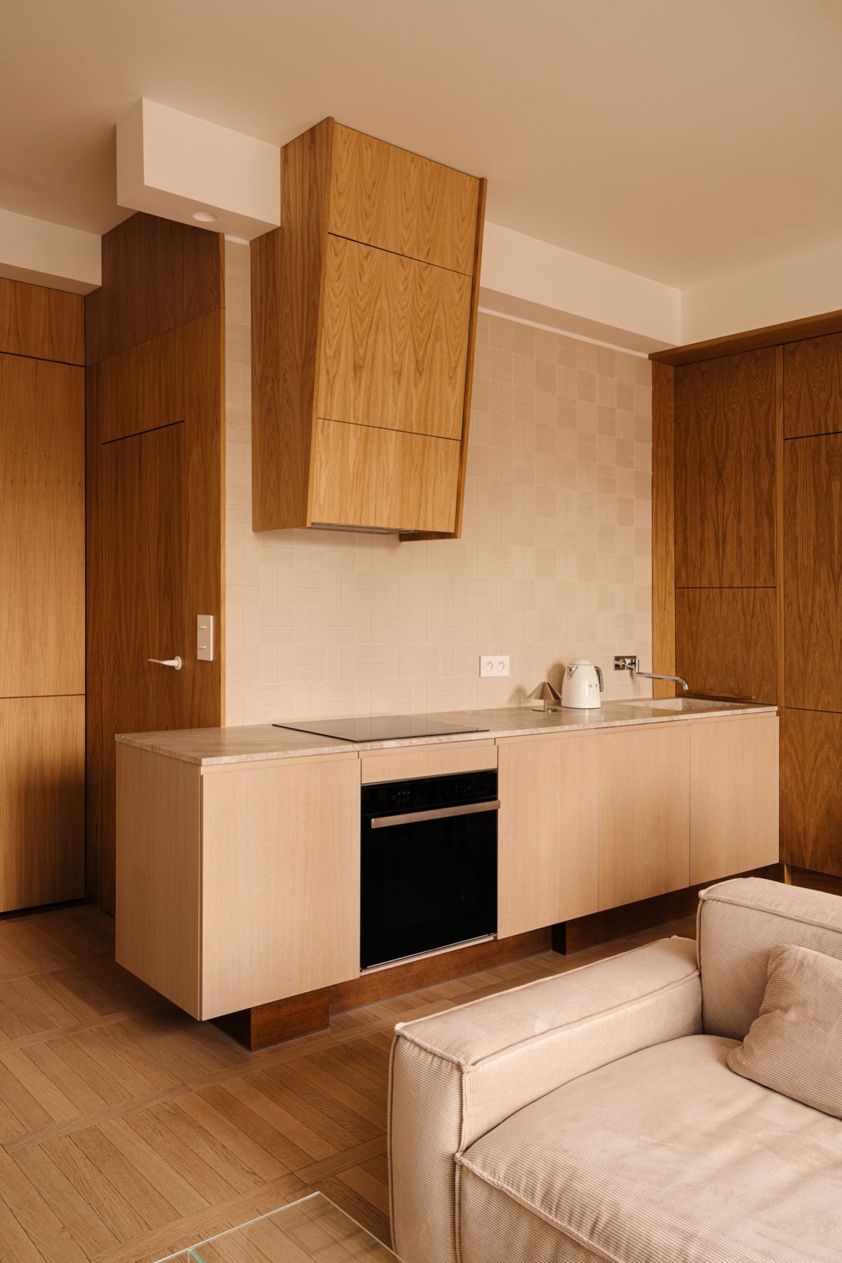 Jasna kuchnia z drewnianymi frontami, minimalistycznym okapem i blatem z kamienia. Widoczny fragment sofy w neutralnym odcieniu. Parkiet w jodełkę.