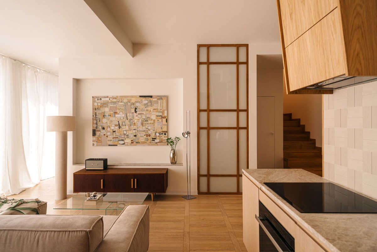 Jasne wnętrze z drewnianymi schodami w tle, widok z perspektywy kuchni z blatem z kamienia. Na ścianie obraz w beżowych tonacjach i drewniana zabudowa.