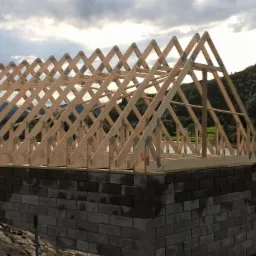 Konstrukcja drewniana dachu w trakcie budowy, widok na ściany z szarego bloczka betonowego i malowniczy krajobraz z jeziorem i zalesionymi wzgórzami w tle.